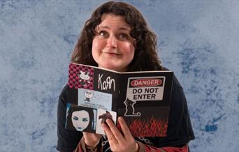 Tiff gazes whimsically at the camera with a diary covered in stickers in her hands. She wears a black t-shirt with long red and grey striped sleeves.