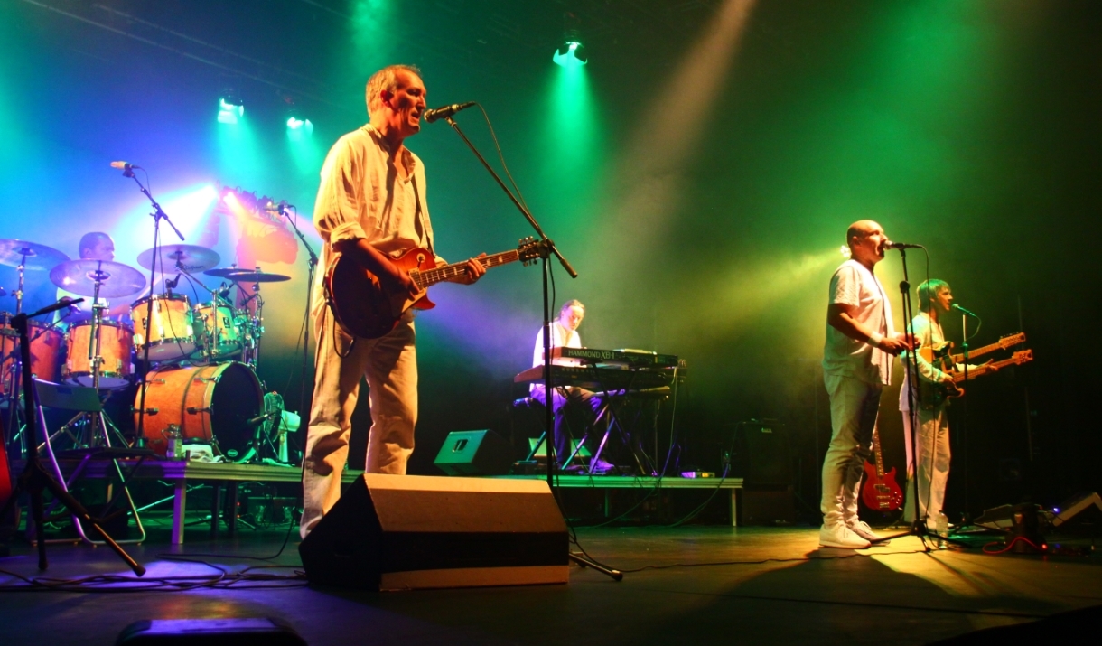 A photo of G2 Definitive Genesis performing on a stage. They are all wearing white clothing. Green, red, white and purple lights illuminate the stage.