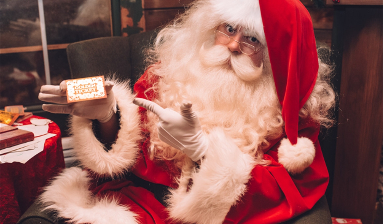 Father Christmas is dressed in his finest plush velvet red and white suit. He is looking at the camera and holding a golden ticket.