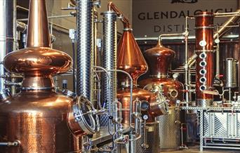 An image from Glendalough distillery from the still house. An assortment of shiny copper stills and stainless steel pipes. In the backgroud is the gle