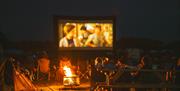 People watching outdoor cinema at night around bonfire