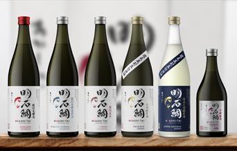 A picture of different sake bottles from Akash-Tai brewery in Japan lined up on a wooden board with a soft cream coloured background.