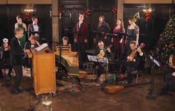 Small orchestra sat round a harpsichord with four singers standing behind them. All stood in a festive panelled room.