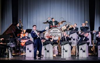 Vintage big band set up with musicians sat behind branded Alex Mendham podiums made of silver and black glitter. All sat in front of a grey curtain. A