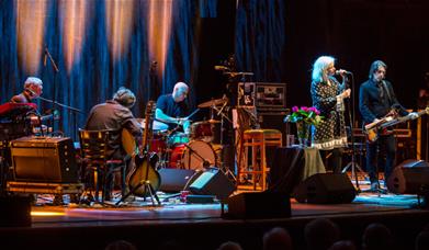 Band performing live on stage under warm lighting, with a female vocalist at the microphone and musicians playing guitar, keyboard, and drums. 