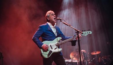 A photo of Graham Gouldman. He is playing an electric guitar while singing