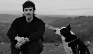 Black and white photo of person crouching outdoors beside a dog looking up.