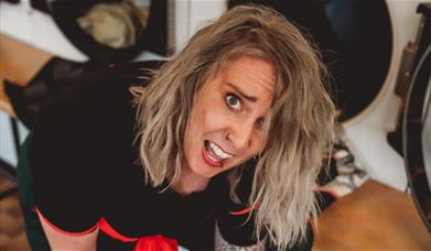 Laura Belbin leaning toward the camera in a laundrette, pulling a playful face. 