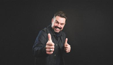 Portrait of Charlie Baker smiling in a dark jacket giving two thumbs up against a black background.