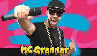 Mc Grammar holding mic toward camera with "MC GRAMMAR" text on a colourful pink and blue background