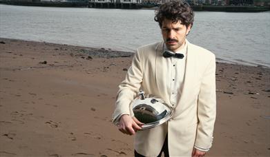 Paddy Young in a white jacket and bow ties holding a silver cloche on a sandy riverbank. 