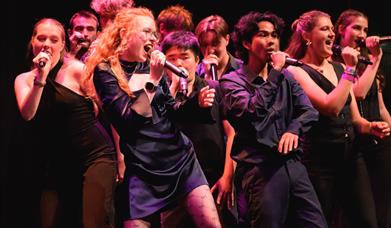 Group of performers singing on stage with microphones under colourful stage lighting. 