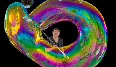 Performer creating a giant rainbow soap bubble against a black background.
