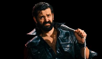 Fin Taylor in a black leather jacket holding a microphone against a dark background.
