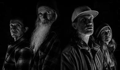 Four people in a dramatic black-and-white portrait against a dark background.