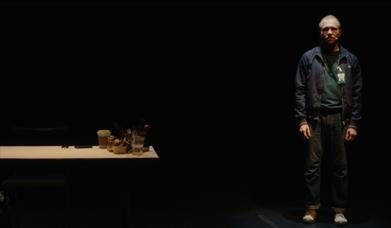 A man stands blankly on a stage, near a table. 