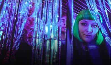 Four people peering through glittering tinsel curtains in purple and teal light.