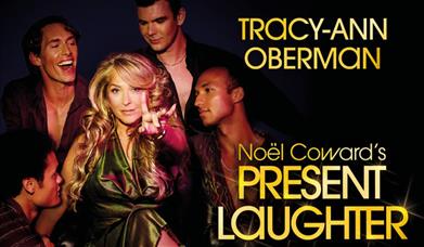 A glamorous woman points at the camera, as 4 young men surround her.