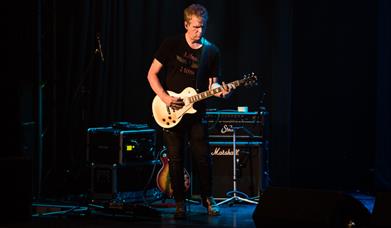 Dion Smith playing a cream Les Paul electric guitar on a darkened stage, with Marshall amps behind him.  