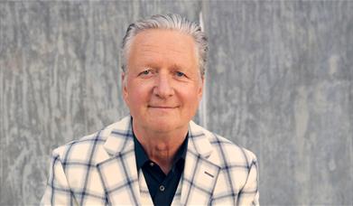 Portrait of a man with grey hair wearing a light checked blazer and dark shirt, standing against a textured grey wall