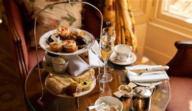 A tiered plate of small cakes and sandwiches along with champagne glasses and tea cups on a small round mahogany table