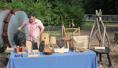 man standing in woodland by table displaying wooden craft objects 