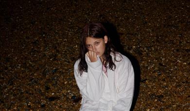 It's evening, a young woman in a white dressing gown sits down on a beach. 