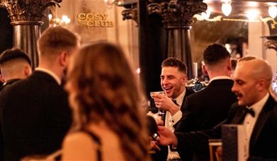 A group of people dressed in black tie attire drinking cocktails in one of Cosy Club's venues. 