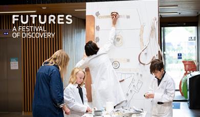 A group of children wearing lab coats, along with a researcher facilitator are crafting a display of urban planning using household items.