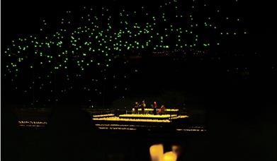 The image shows an outdoor area with hundreds of drones in the sky and a string quartet playing surrounded by candles.