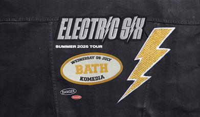 Jacket with patches: "Electric Six," "Summer 2025 Tour," "Wednesday 09 July BATH Komedia," and a lightning bolt and danger sign.