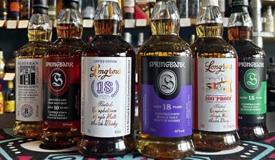 6 bottles of whisky sitting on a countertop with a wall  of spirits behind the,. The whiskies are Springbank 10 Amontillado 55%, Springbank 15 46%, Sp