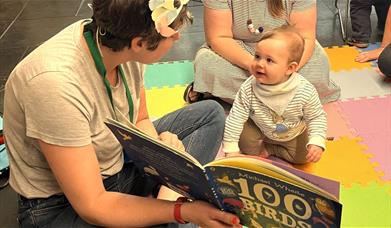 A woman reads a picture book to a baby, who is sat on a soft coloured mat