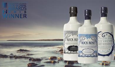 An image of a misty rocky beach off the coast of scotland. The image has 3 bottles of rock rose gin on it. Ceramic white bottles with blue writingg