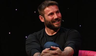 A smiling Ben Cohen in a black t-shirt speaking on a darkened stage.