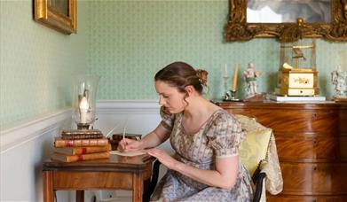 A woman in Regency clothing writing at a writing desk