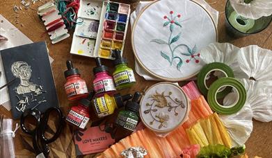 A colourful chaos of crafting tools and materials with a Regency theme sprawled out including lino tools, scissors, threads, embroidery hoops, paints 