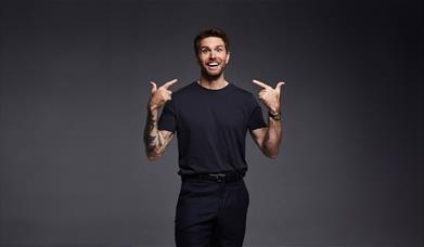 Joel Dommett in a black T-shirt pointing at his face with both hands