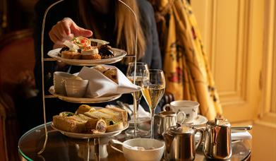Cream tea on tiered plates on a dark wood table with two glasses of champagne and a lady eating. 