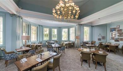 A horizontal view of the restaurant with soft chairs, wooden tables and a chandelier hanging above.
