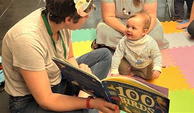 A woman reads a story to a baby sitting on a colourful puzzle floor
