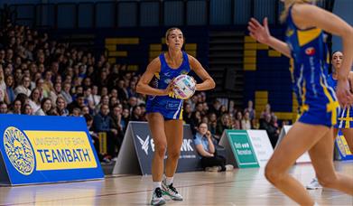 A Team Bath Netball player, on court with ball in hand, lines up a pass as a large crowd watches on