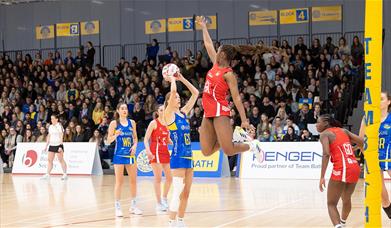 A defender, wearing red, leaps into the air as a Team Bath Netball shooter, wearing blue, lines up a shot. There is a stand of spectators watching beh