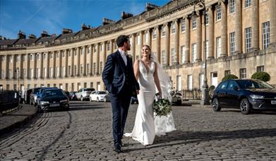 A bride and groom holding hands and walking along the Royal Crescent in Bath