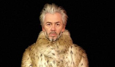 A man stands in front of a black background, he wears a large animalistic coat.