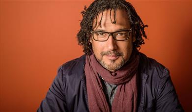 Image of Professor David Olusoga wearing a dark jacket and maroon scarf, is seated against a warm, reddish-orange backdrop
