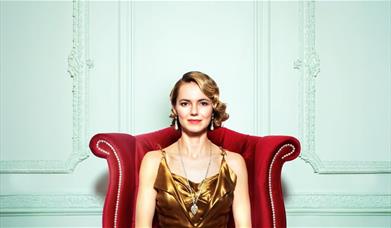 A woman in 1920s attire is sat down on a red chair, she's smiling. 