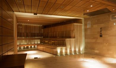 A large glass fronted sauna with wooden staged steps to sit on and low warm uplighters. 