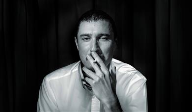 A black and white photo of the comedian Tom Rosenthal. He's smoking a Cigarette 