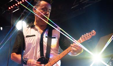 A guitarist plays on stage surrounded by bright stage lights